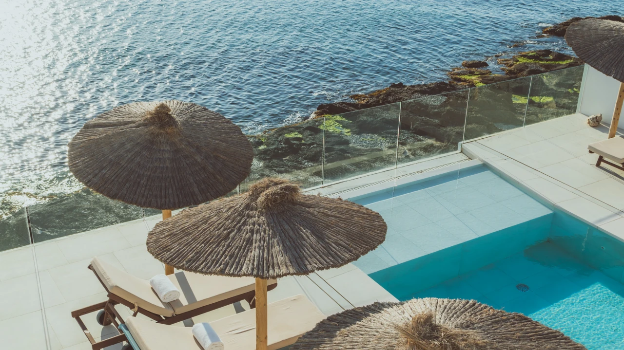 Piscina junto al mar con sombrillas de paja y tumbonas.