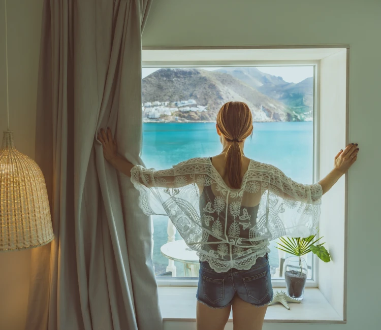 Mujer mirando el mar por una ventana, vistiendo blusa de encaje y shorts.