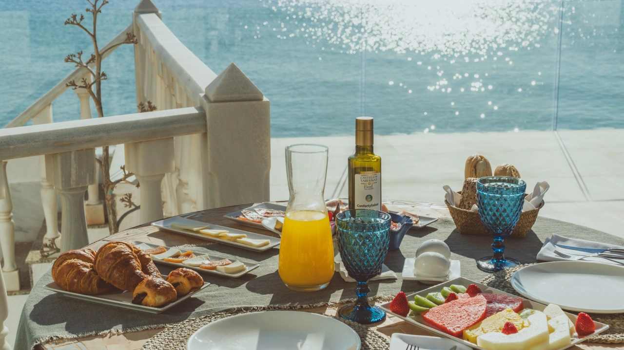 Desayuno con vista al mar: fruta, croissants y jugo de naranja en mesa.