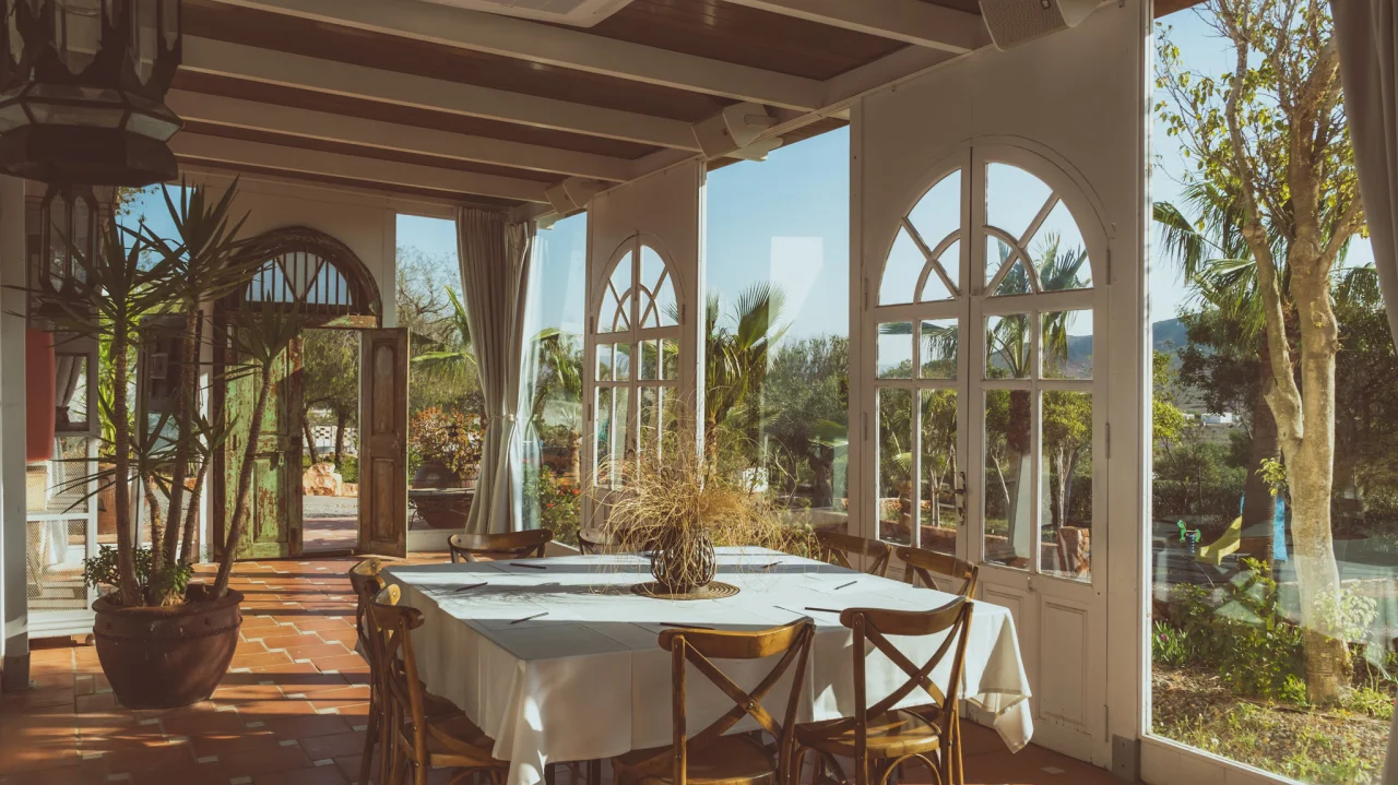 Terraza iluminada con mesa y sillas de madera, rodeada de plantas y ventanas.