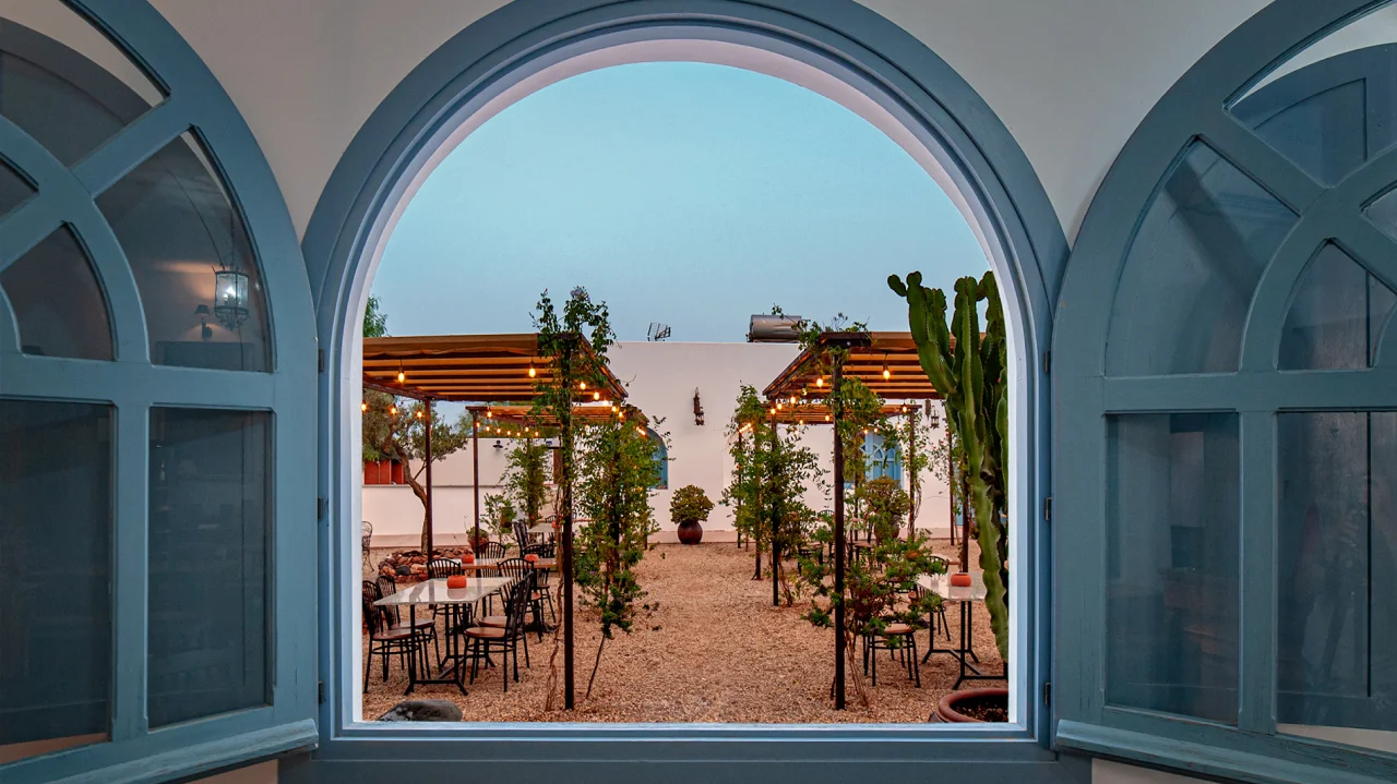 Vista de un patio con mesas, plantas y p&eacute;rgolas iluminadas desde ventana arqueada.