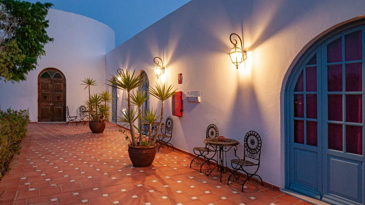 Pasillo exterior iluminado con mesas, sillas y plantas en macetas.