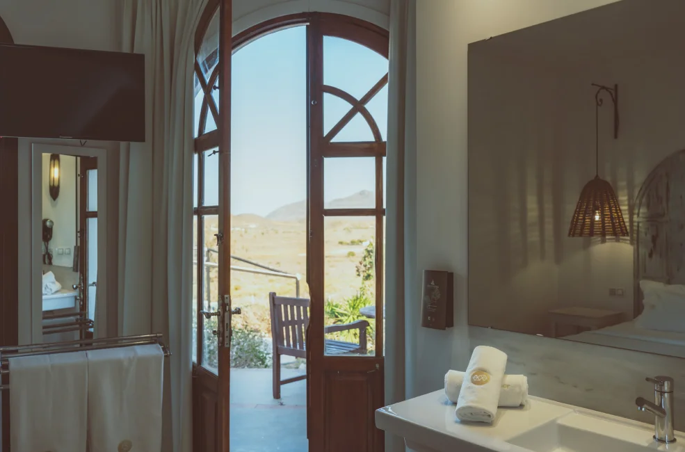 Ba&ntilde;o con puerta abierta al campo, toallas en el lavabo y espejo reflectante.