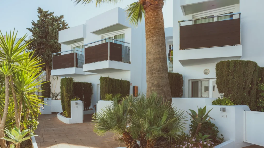 Edificio moderno con balcones y jard&iacute;n, rodeado de palmeras y arbustos.
