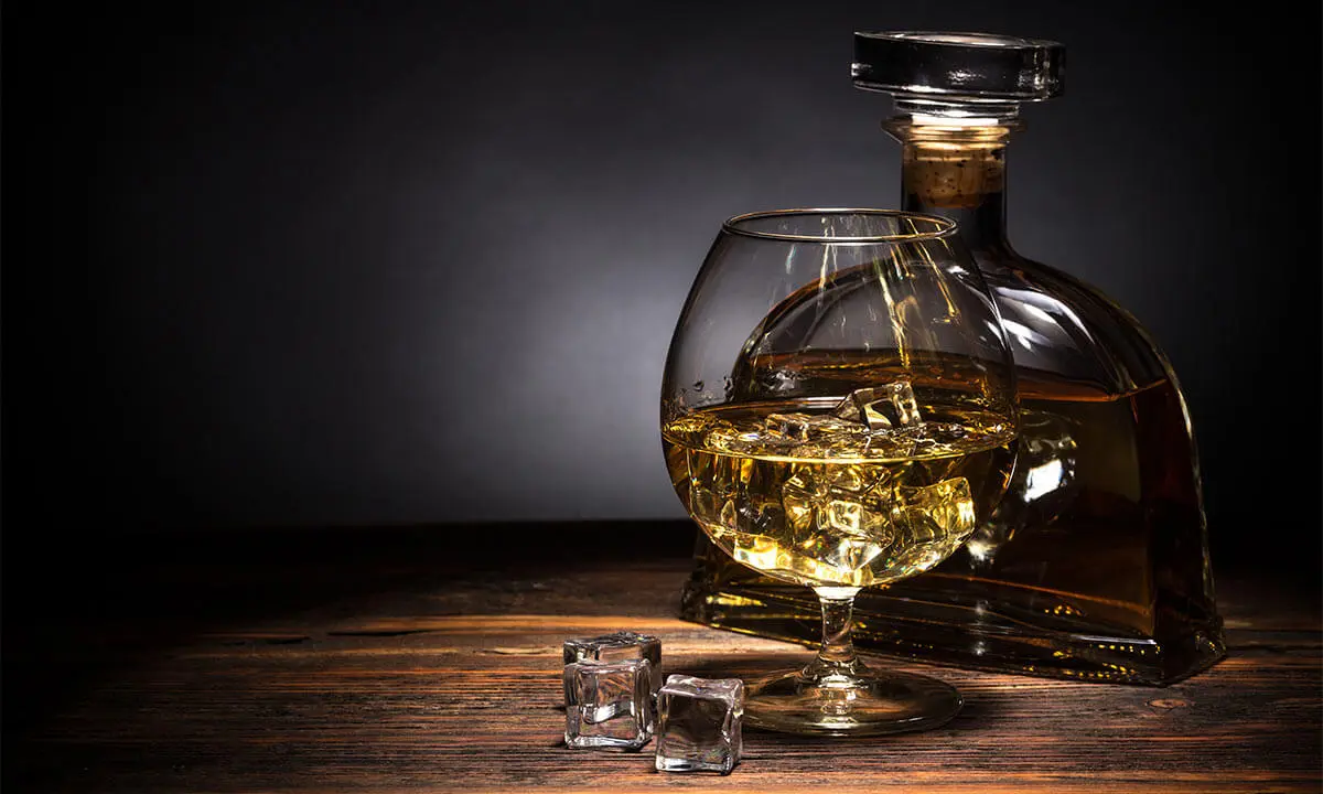 Botella con licor dorado y copa, sobre mesa de madera oscura, fondo difuso.