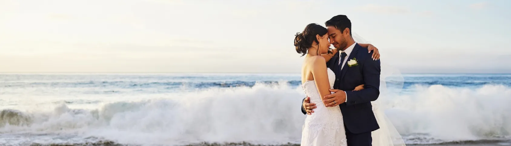 Pareja de reci&eacute;n casados bes&aacute;ndose junto al mar, con espuma de olas.