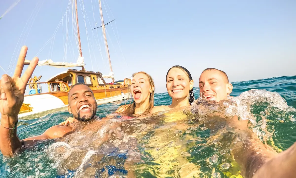 Grupo de amigos nadando en el mar y saludando junto a un barco.