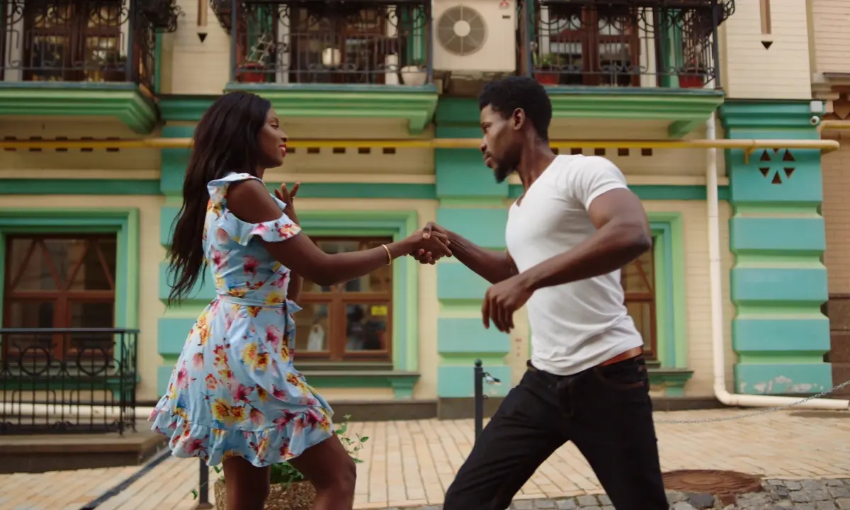 Pareja bailando frente a edificios coloridos, tom&aacute;ndose de las manos
