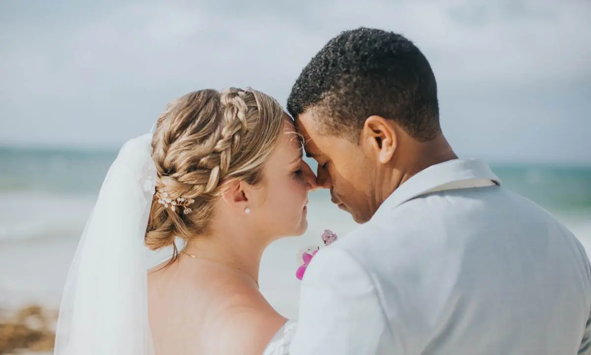 Pareja de novios bes&aacute;ndose en la playa, con traje blanco y cielo nublado