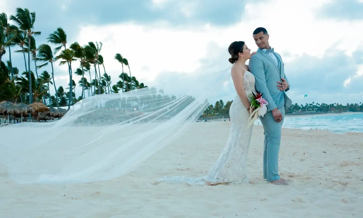 Pareja en boda bajo palmeras en la playa, posando con vestido y traje azul