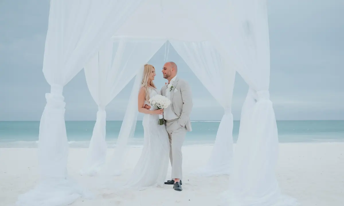 Pareja de novios con ramo, bajo una estructura de tela blanca junto al mar