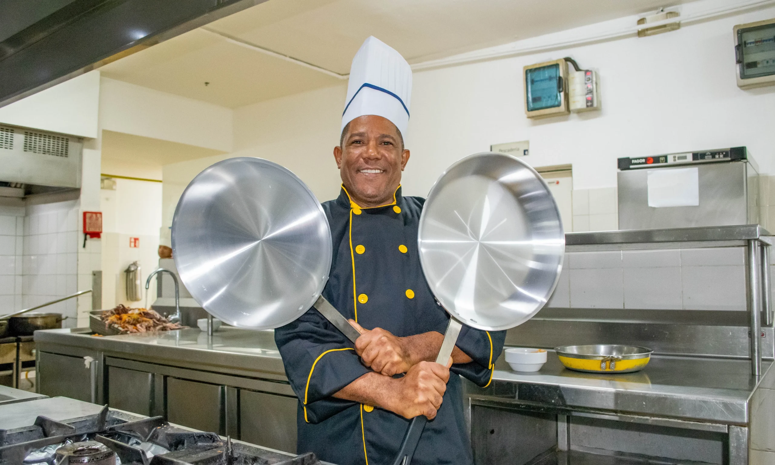 Chef en cocina industrial sonriente con dos sartenes met&aacute;licas al frente