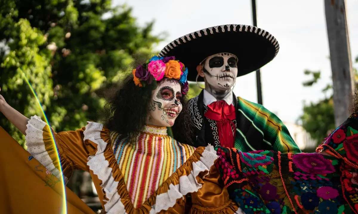 Pareja de calaveras con trajes coloridos, sombrero y flores en un desfile.