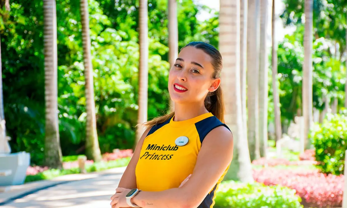 Mujer con uniforme amarillo y azul, sonriendo en un jard&iacute;n entre columnas verdes
