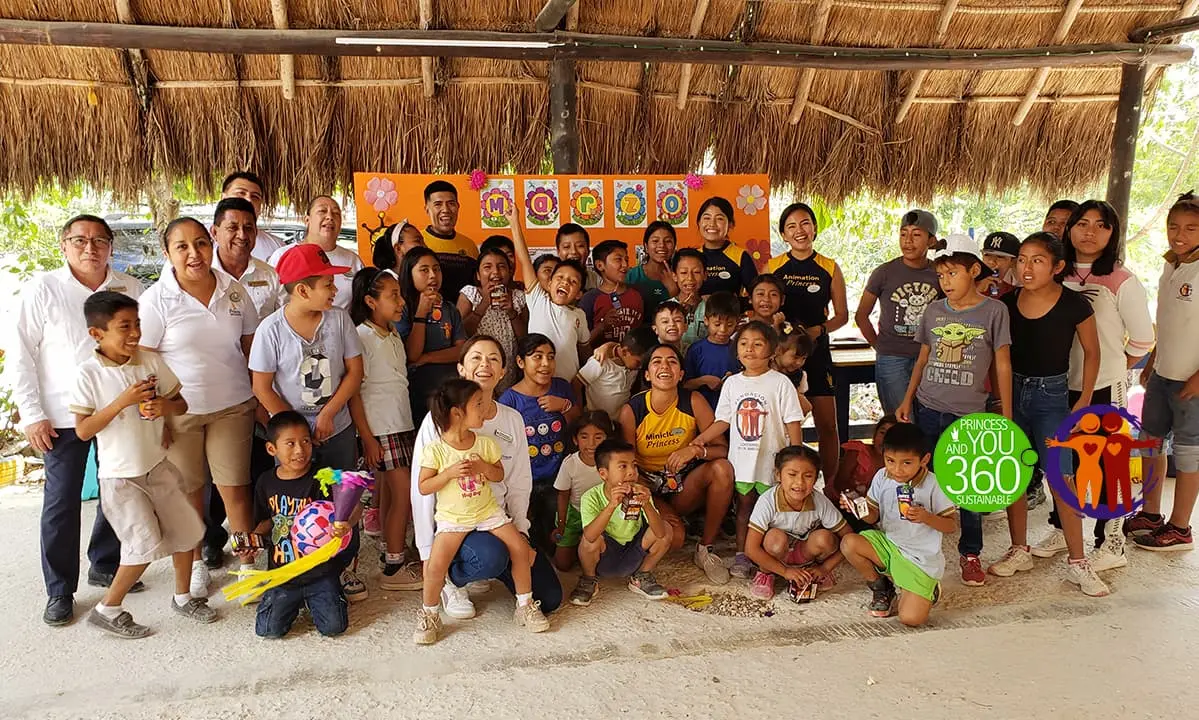 Grupo de personas y ni&ntilde;os posan bajo techo de paja con pancarta infantil.