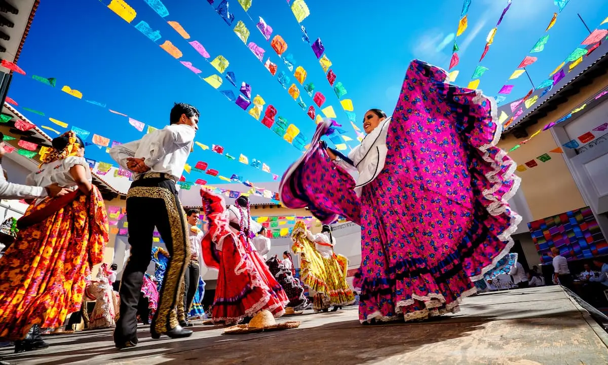 Bailes con trajes coloridos bajo cielo azul y banderines en una fiesta tradicional.