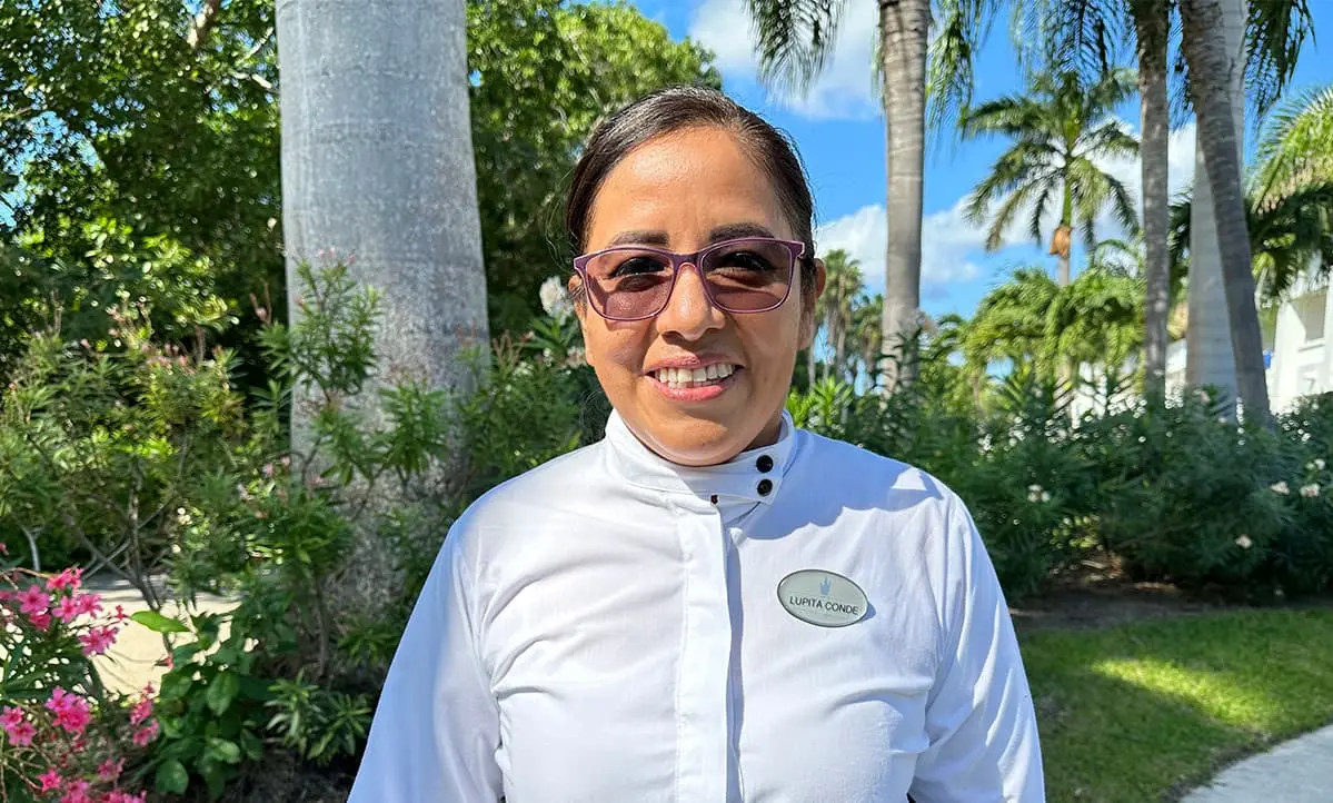 Mujer con uniforme blanco y gafas, sonriendo en jard&iacute;n con palmeras al fondo
