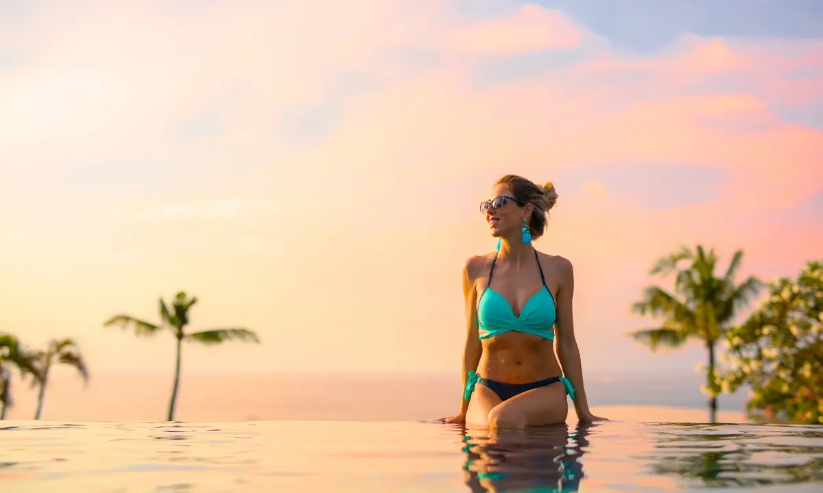 Mujer con bikini turquesa y gafas, de pie en una piscina al atardecer.