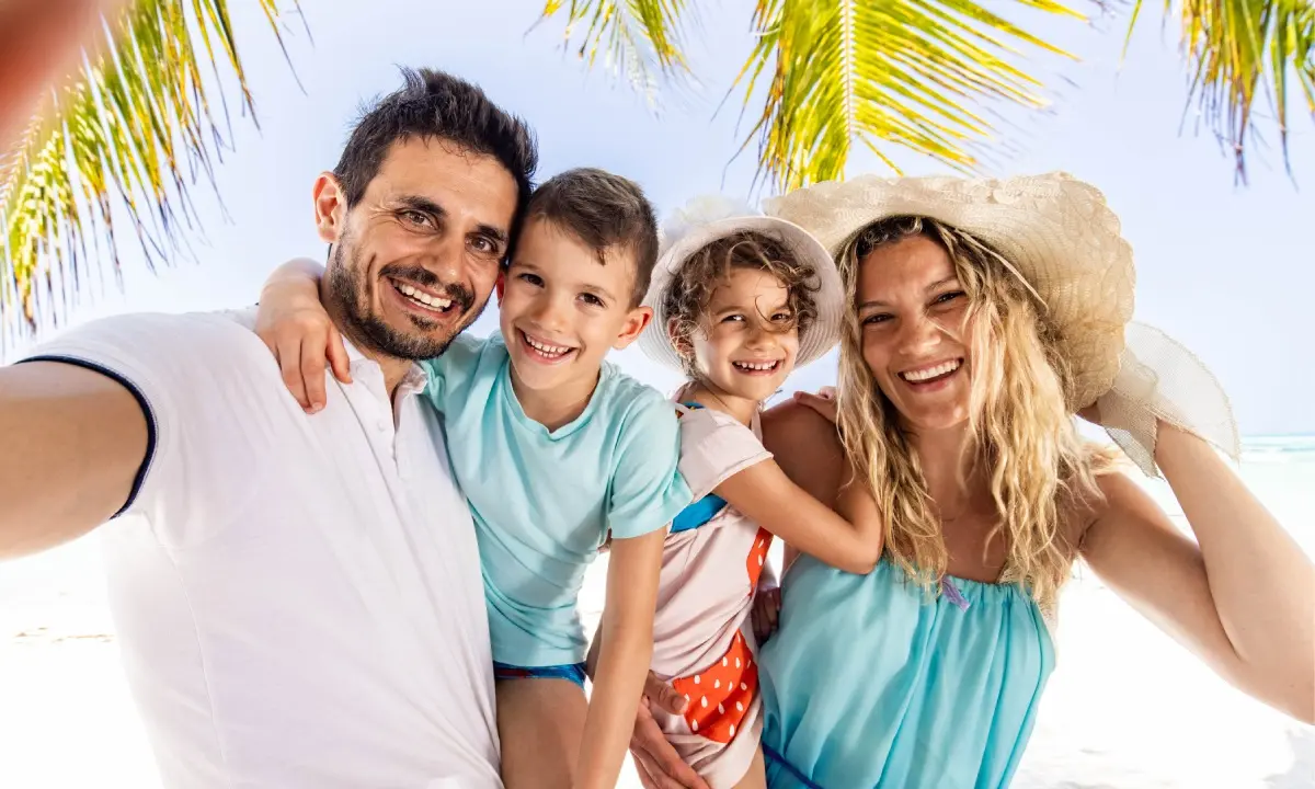 Familia sonriente en la playa con sombreros de paja y palmeras al sol.