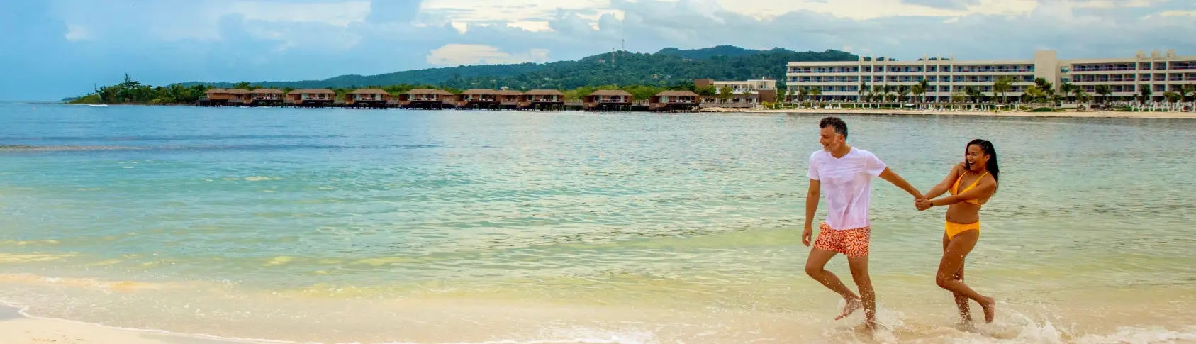 Pareja camina en la orilla de la playa, con resort y monta&ntilde;as al fondo
