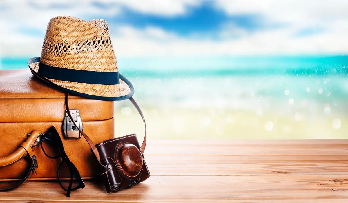 Sombrero de paja sobre ba&uacute;l de cuero junto a playa azul, con espacio para texto