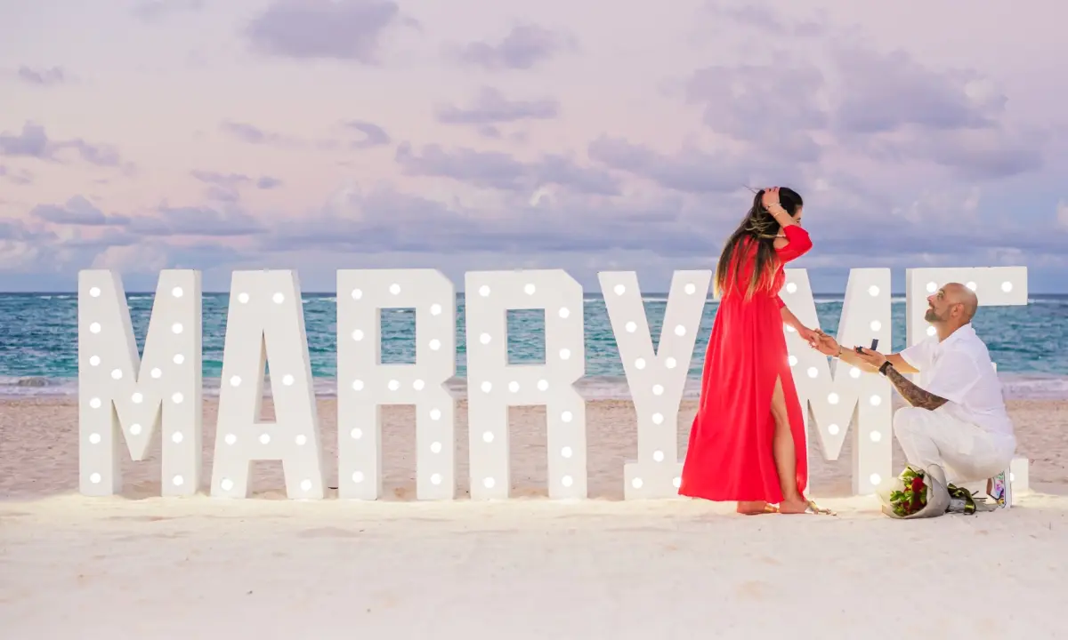 Pareja en la playa, mujer con vestido rojo y hombre arrodillado junto a letras luminosas &ldquo;MARRY ME&rdquo;