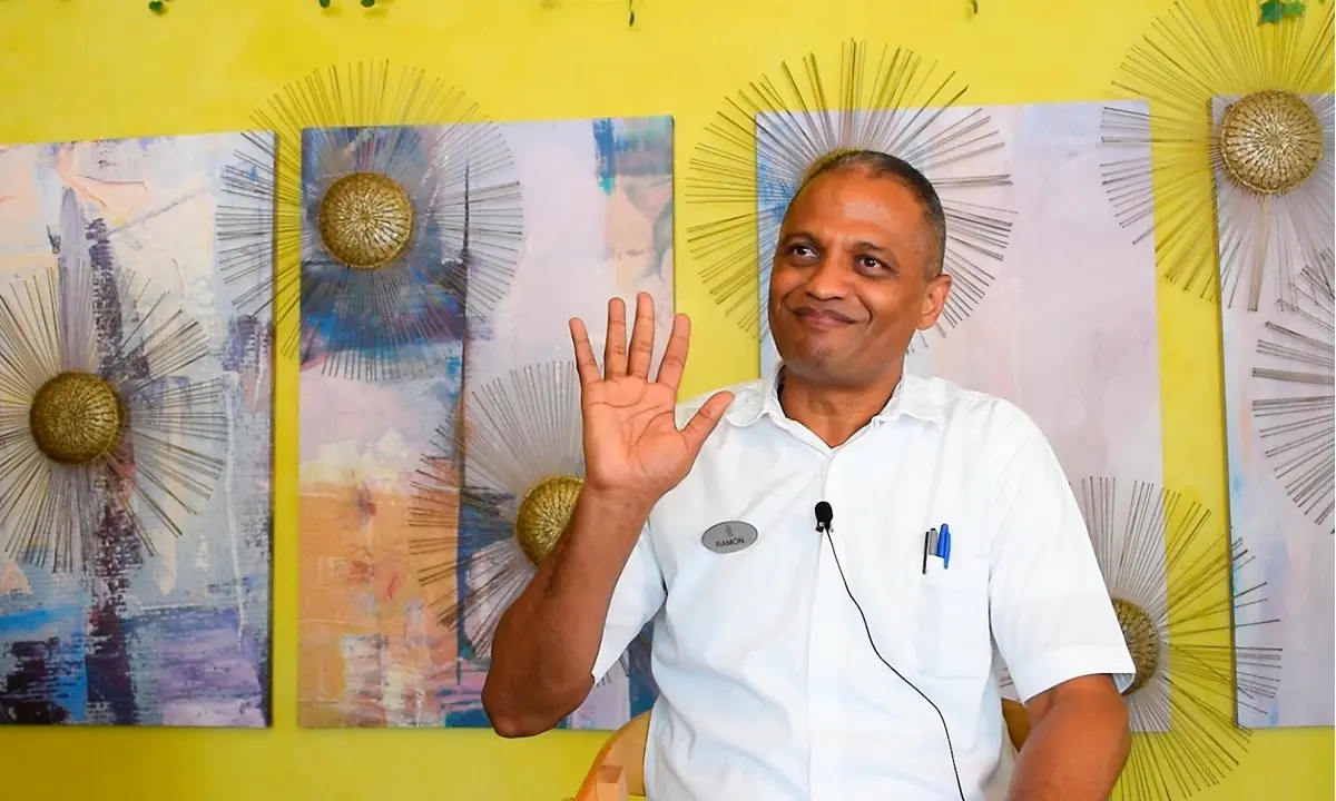 Hombre sonriendo con traje blanco y mano en alto, fondo amarillo decorado