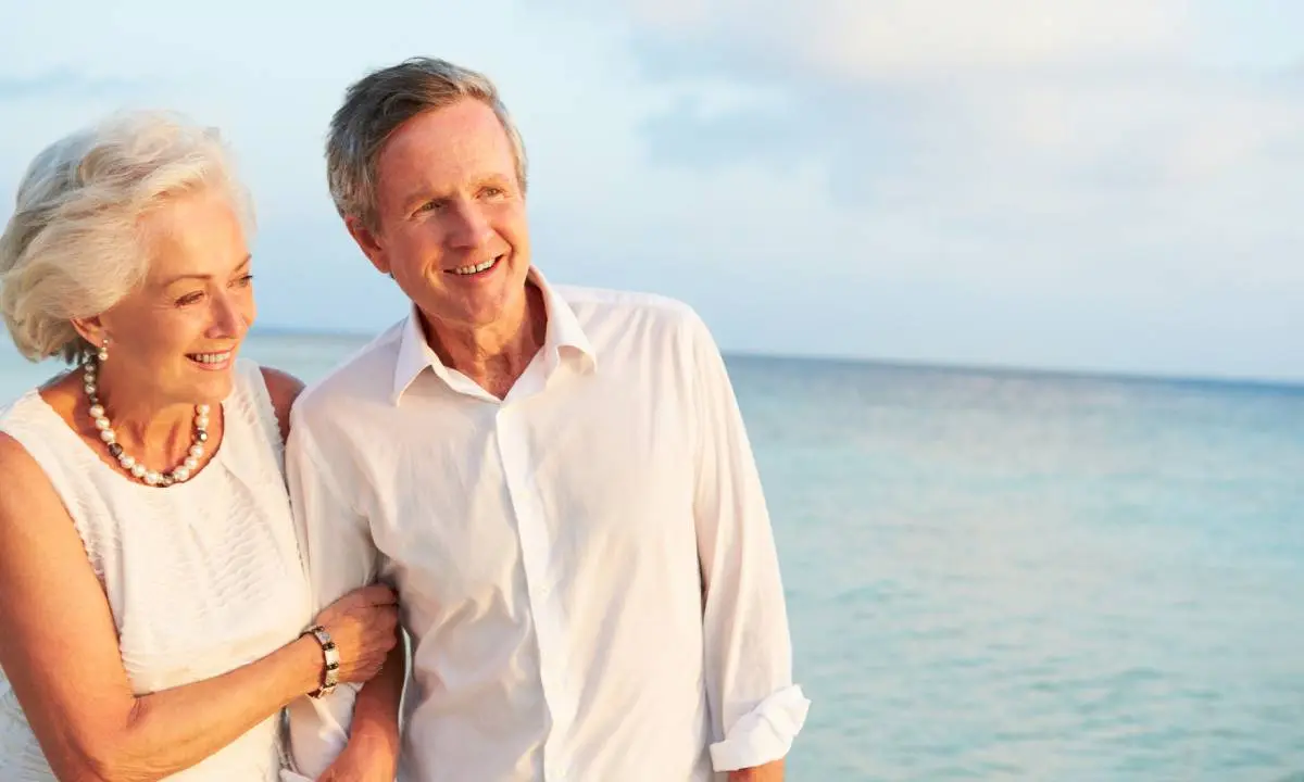 Pareja de adultos mayores sonrientes junto al mar, con ropa blanca.