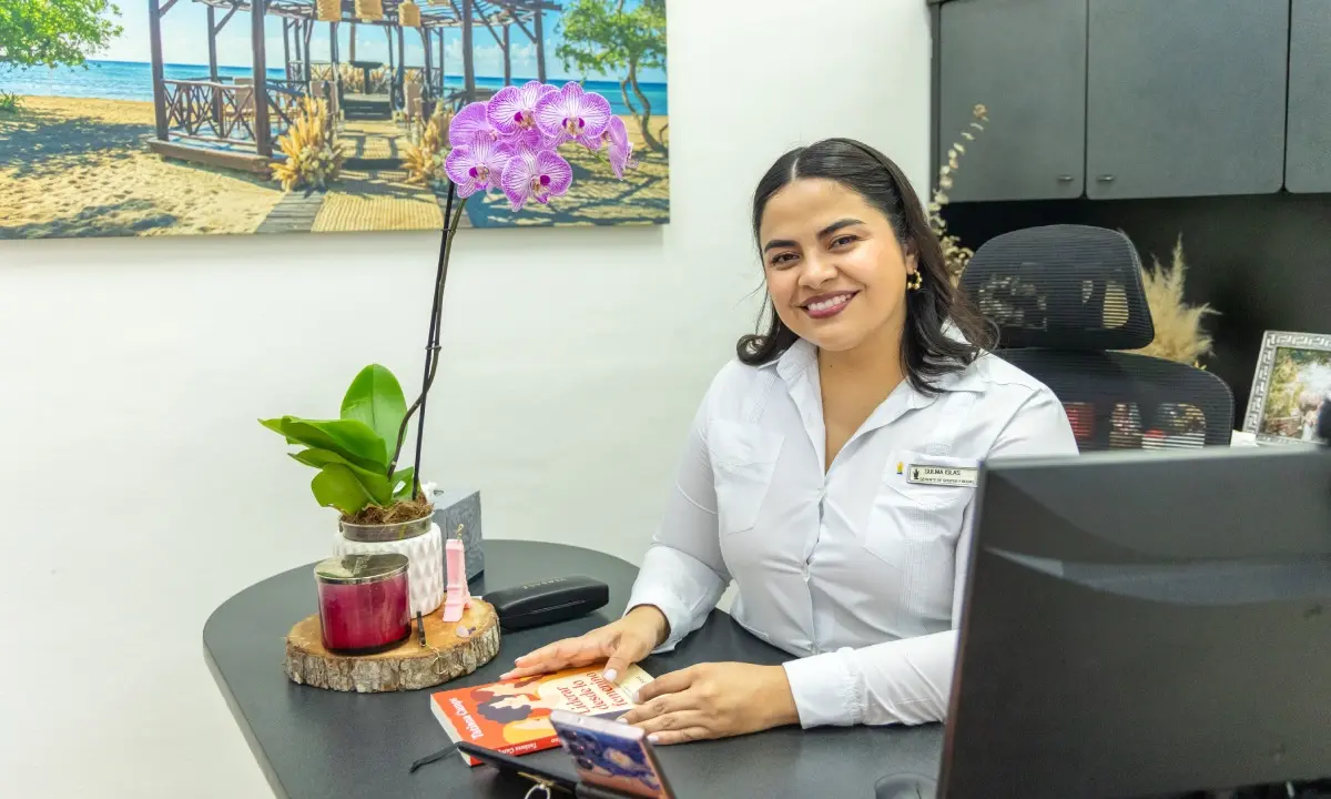 Mujer sonriente atiende en recepci&oacute;n, con escritorio y decoraci&oacute;n floral morada.