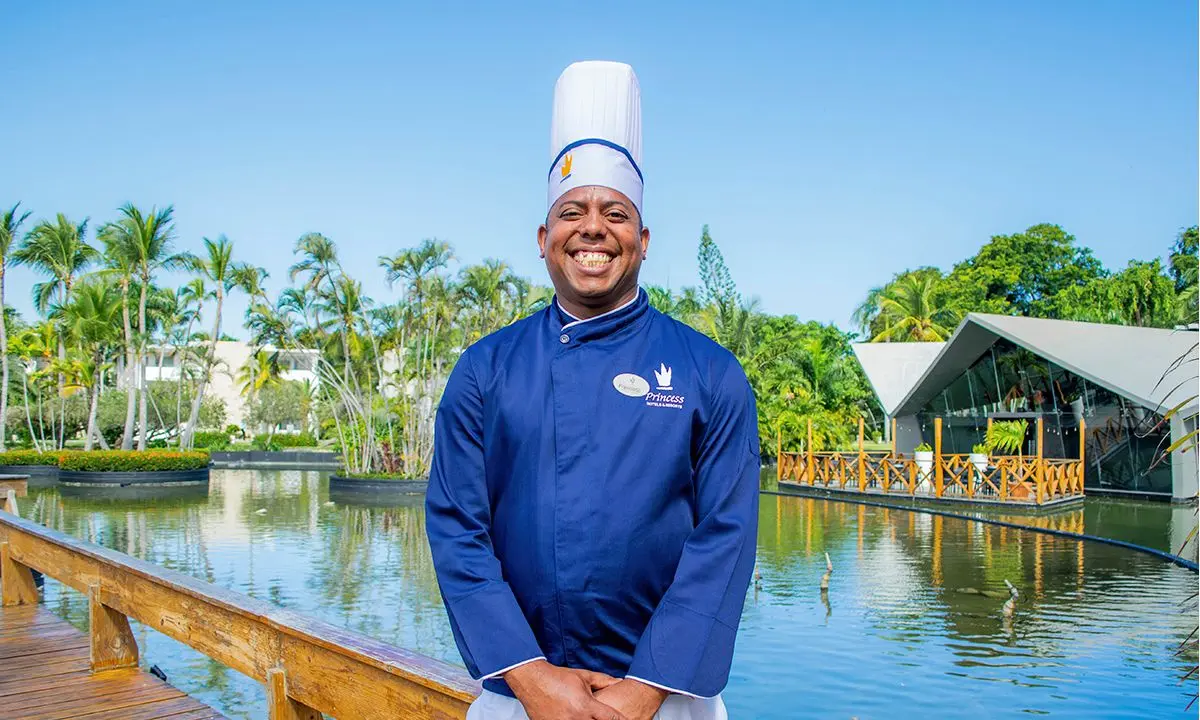 Chef con uniforme azul y gorro blanco, sonriendo junto a un estanque con palmeras