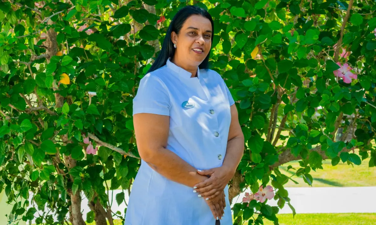 Mujer chef con uniforme blanco, sonr&iacute;e entre &aacute;rboles verdes y flores.
