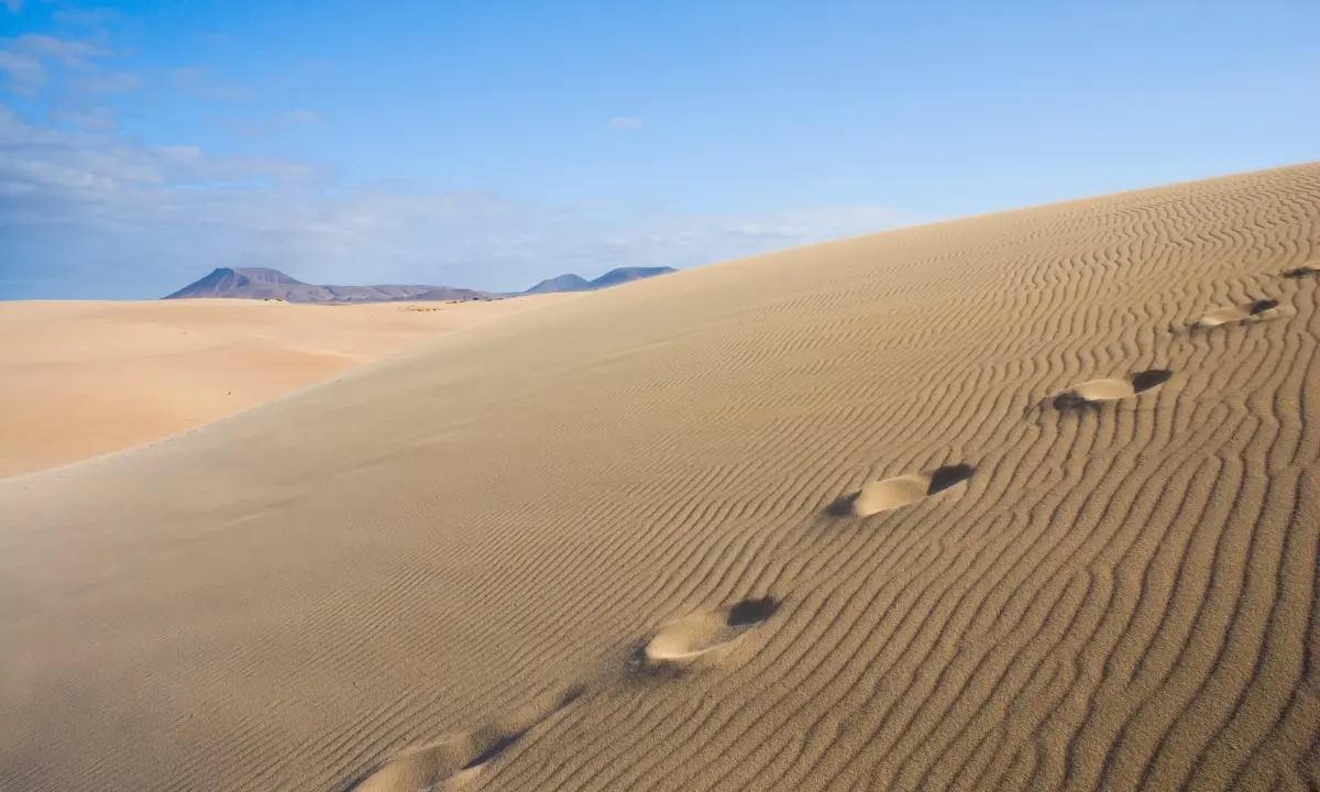 Dunas de arena con huellas y monta&ntilde;as al fondo bajo cielo despejado