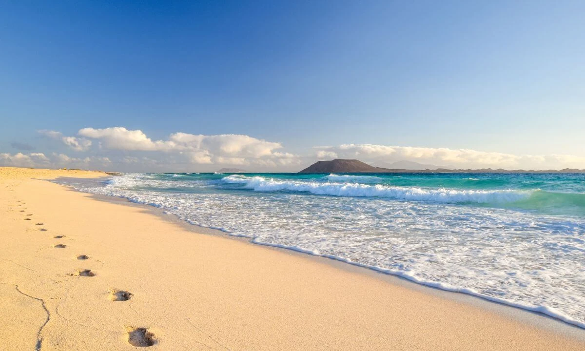Playa de arena dorada con olas azules y un volc&aacute;n al fondo bajo el cielo claro