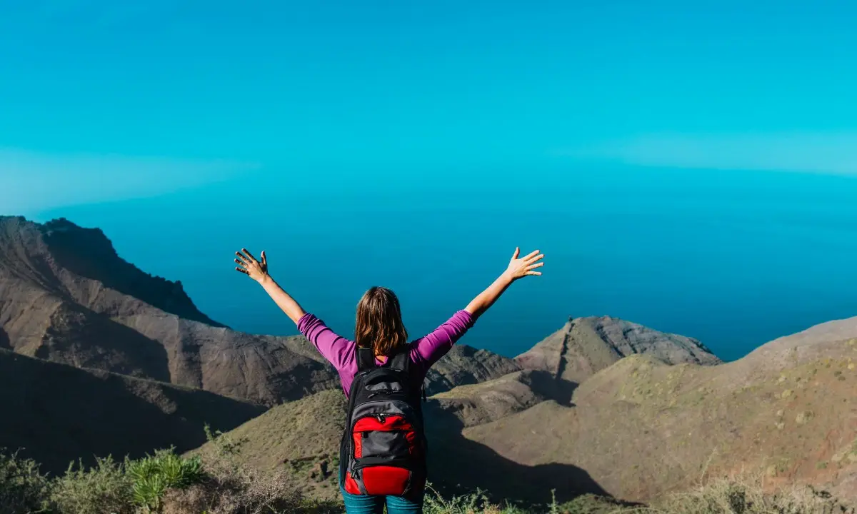 Mujer con mochila alza los brazos frente a monta&ntilde;as y cielo azul