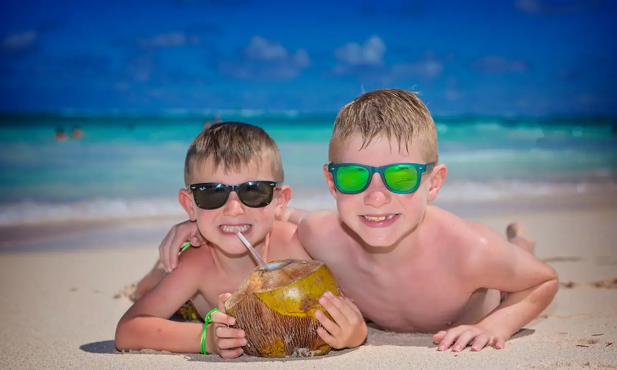 Dos ni&ntilde;os en la playa con gafas y cocos, sonriendo al sol