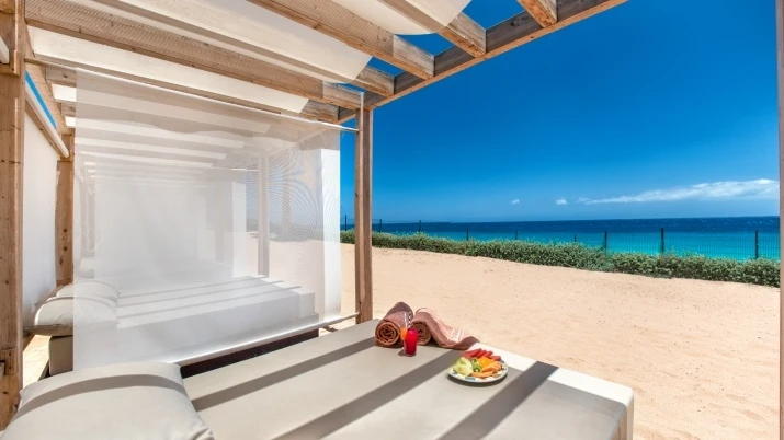Terraza junto al mar con banco blanco y mesa con frutas y toalla.