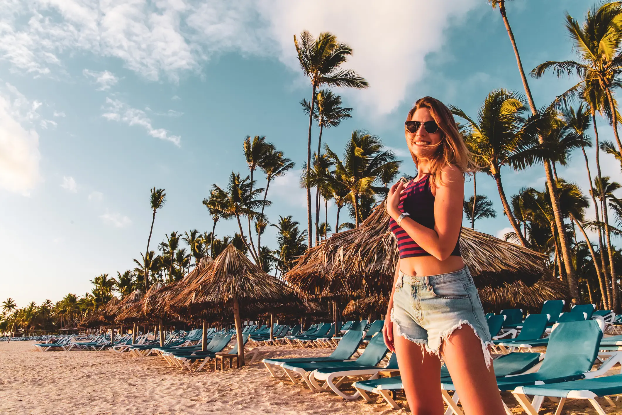 Mujer sonriente con gafas en playa tropical, palmeras y hamacas, sol.