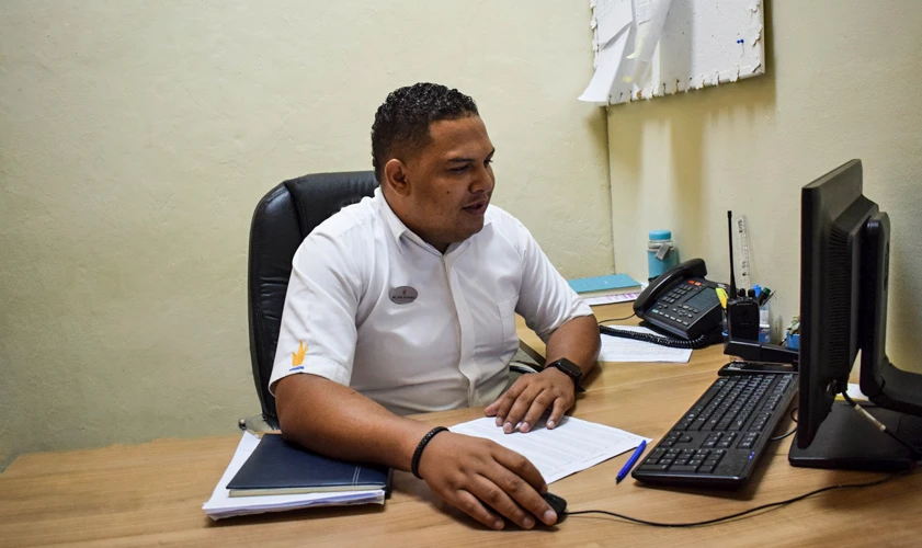 Hombre en oficina revisa documentos frente a computadora y tel&eacute;fono.