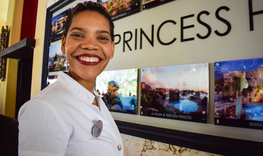 Mujer con uniforme blanco sonriente frente a un cartel que dice &ldquo;PRINCESS&rdquo;