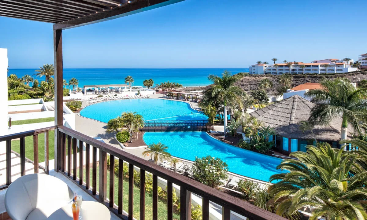 Vista de balc&oacute;n con piscina azul y mar Caribe al fondo, rodeado de palmeras.