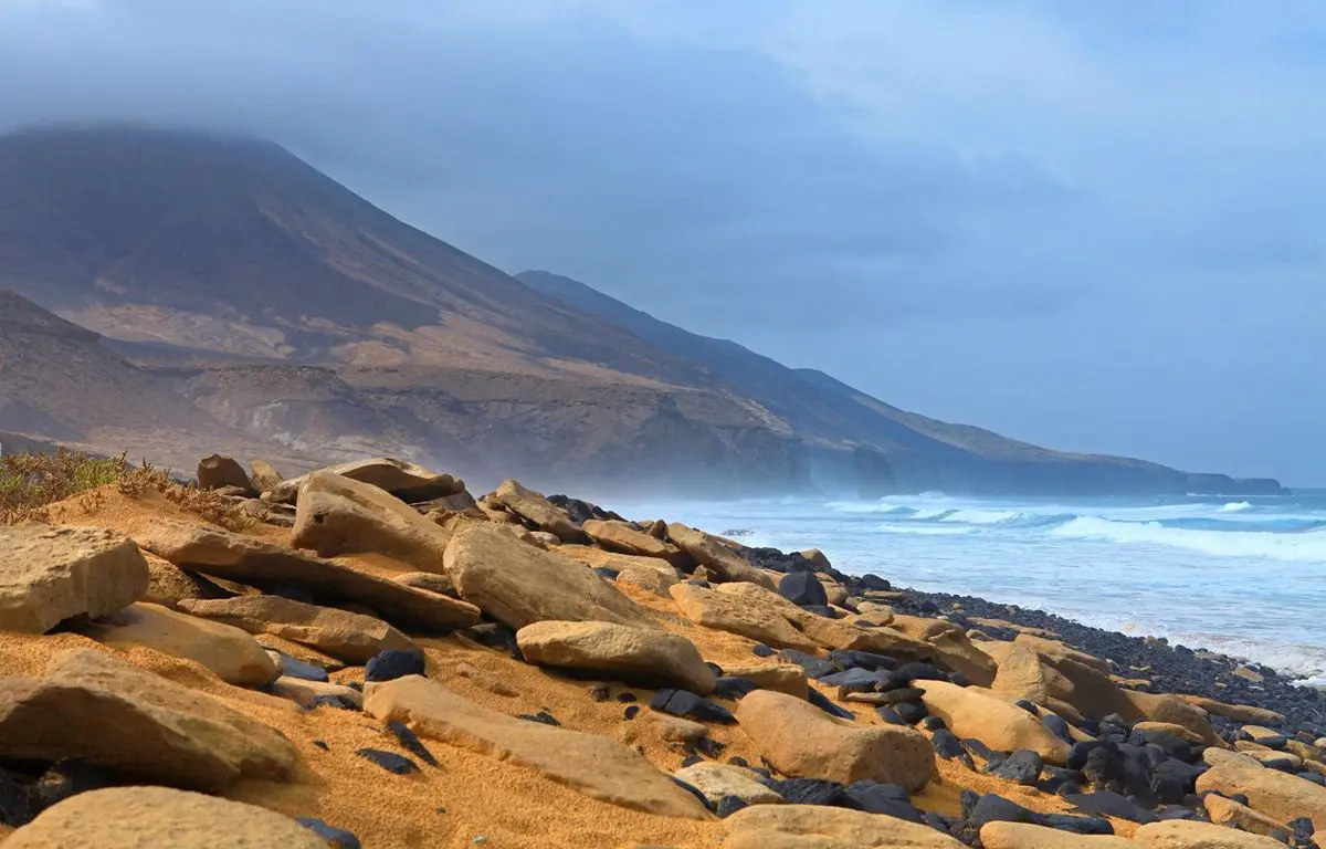 Playa rocosa con monta&ntilde;as al fondo y olas en la costa bajo cielo nublado