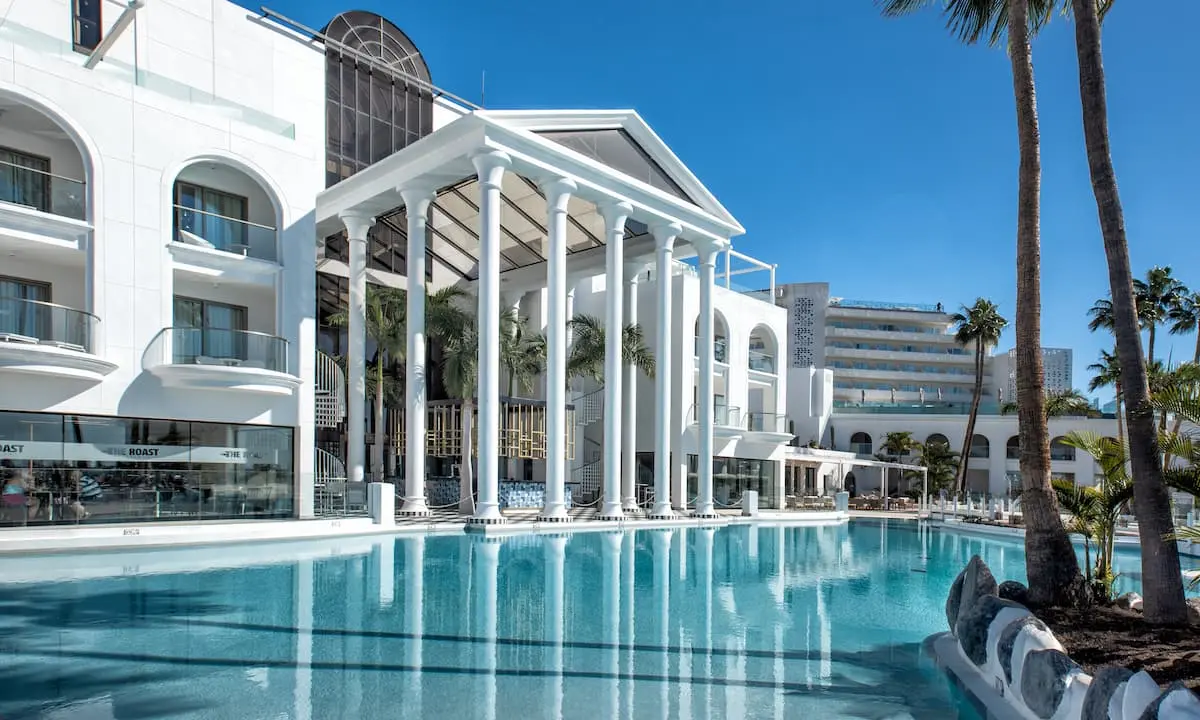 Piscina frente a hotel moderno blanco con columnas y palmeras al cielo azul