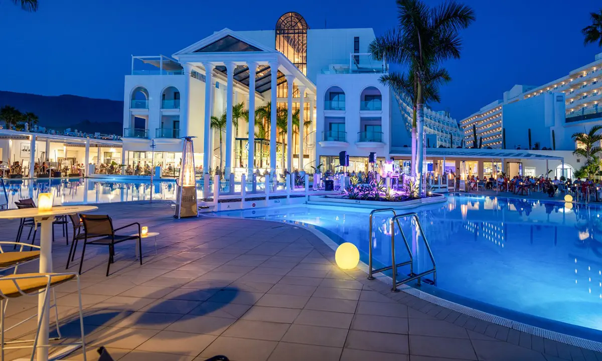Piscina iluminada de hotel con edificio blanco y palmeras al atardecer.