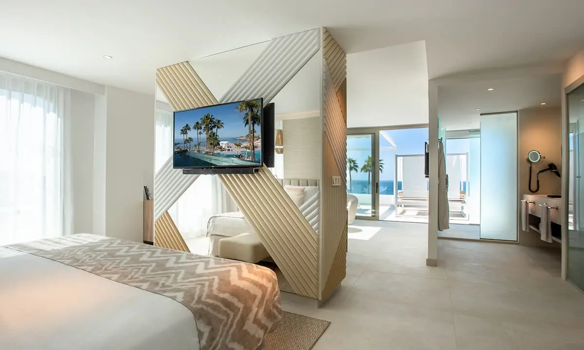 Habitaci&oacute;n moderna con cama, TV y vista al mar por ventanales.