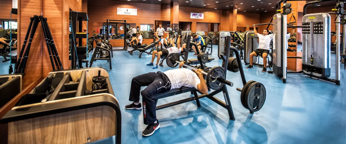 Gimnasio con personas entrenando en m&aacute;quinas y banco con pesas.