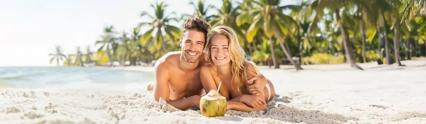 Pareja sonriendo en la playa, acostados en la arena con coco y sombrilla