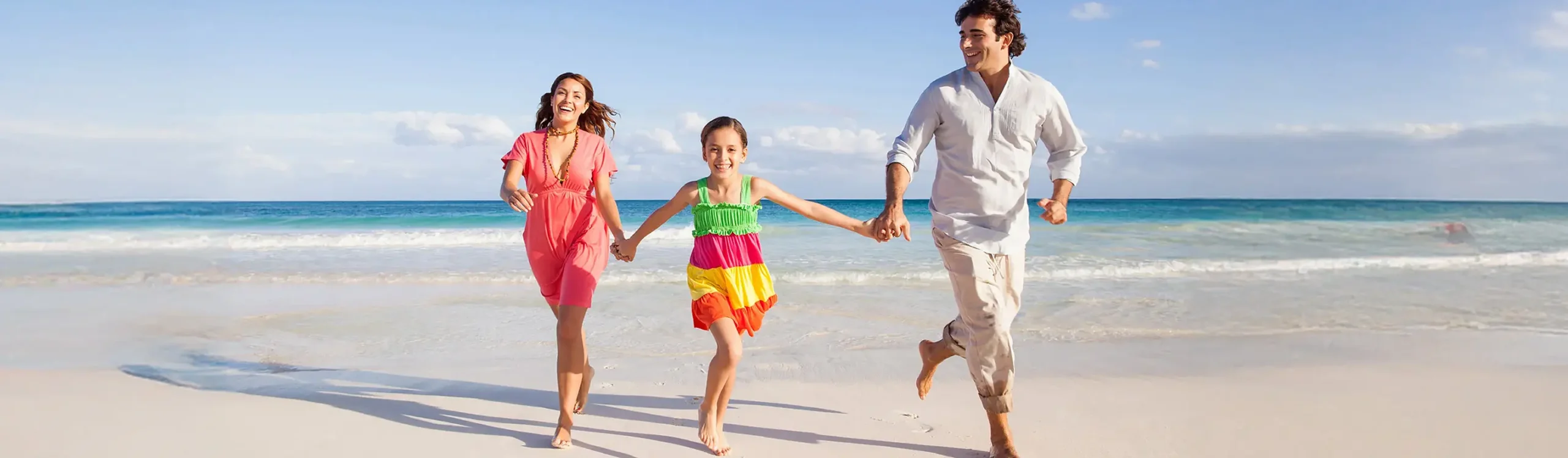 Familia corriendo descalza en la playa, con el cielo despejado y mar al fondo.
