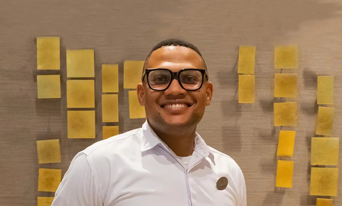Hombre sonriendo con gafas y camisa blanca frente a mosaicos amarillos.