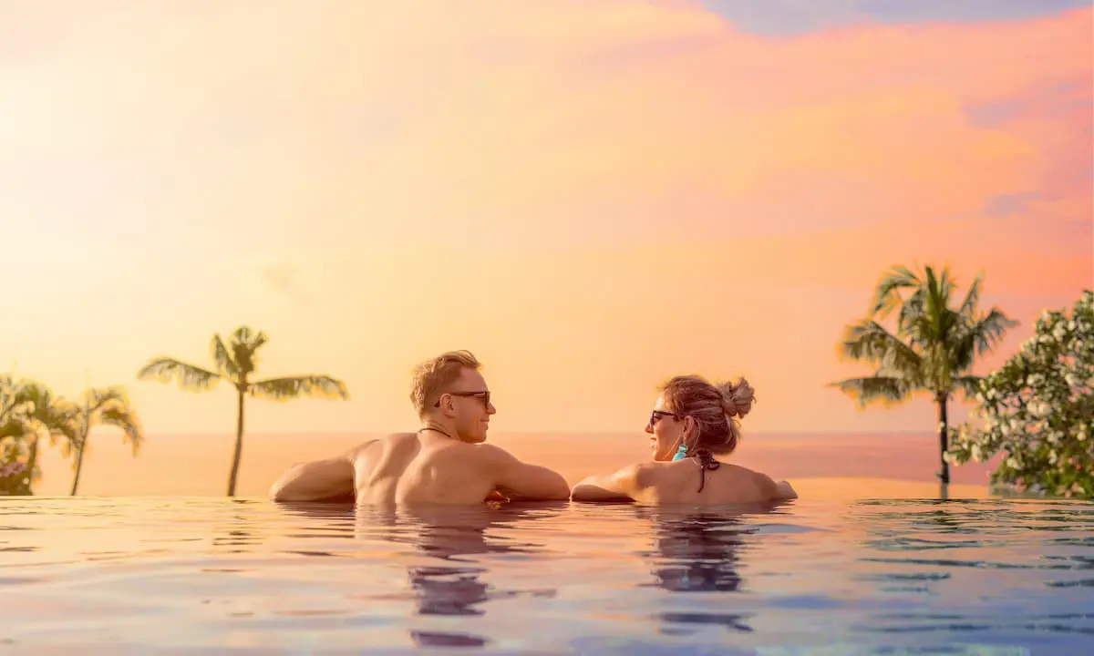 Pareja nadando en una piscina al atardecer, con palmeras y cielo naranja