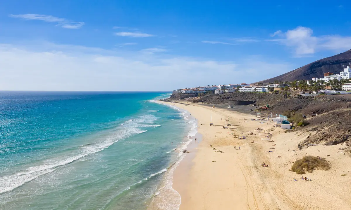 Playa de arena dorada con oleaje y casas blancas al pie de un acantilado